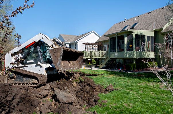 A small digger drops dirt removed from an excavation in back of a suburban house.